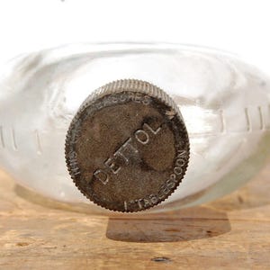 May include: A clear glass bottle with a metal cap. The cap is dark brown and has the word "DETTOL" printed on it. The bottle has a rounded shape and is sitting on a wooden surface.