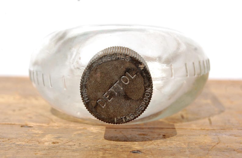 May include: A clear glass bottle with a metal cap. The cap is dark brown and has the word "DETTOL" printed on it. The bottle has a rounded shape and is sitting on a wooden surface.