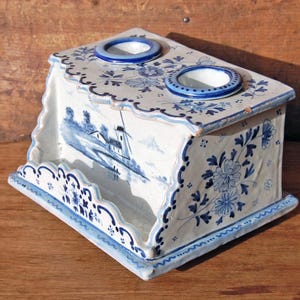 Könnte beinhalten: Eine weiß-blaue Keramikbox mit zwei kreisförmigen Öffnungen oben. Die Box zeigt eine holländische Windmühlen-Szene und florale Muster. Die Kanten sind gewellt und mit blauen Akzenten versehen. Es handelt sich wahrscheinlich um einen dekorativen Gegenstand.