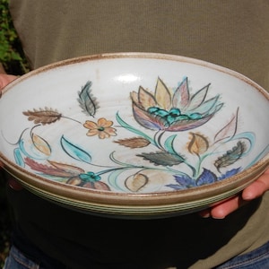 May include: A white ceramic bowl with a floral design in shades of brown, blue, green, and yellow. The bowl has a slightly raised rim and a glazed finish.