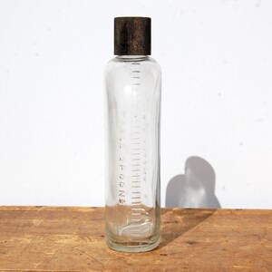 May include: A clear glass bottle with a dark brown cap. The bottle has measurement markings in teaspoons along the side. The bottle is sitting on a wooden surface.