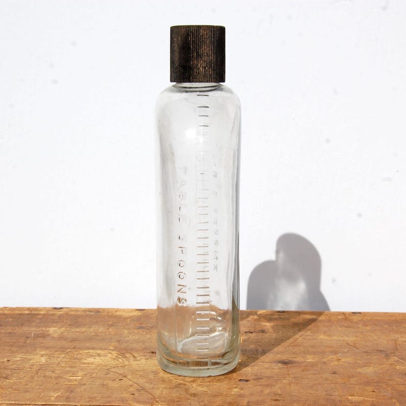 May include: A clear glass bottle with a dark brown cap. The bottle has measurement markings in teaspoons along the side. The bottle is sitting on a wooden surface.