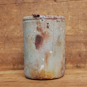 May include: A cylindrical ceramic container with a textured, vertically ribbed surface. The container has a light green and tan color with reddish-brown accents. The top edge has a rough, uneven texture with small, dark brown pieces.