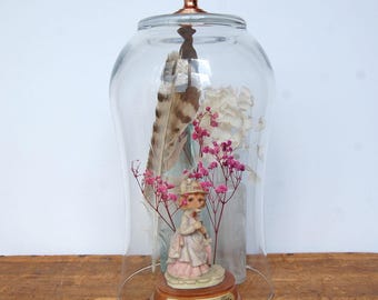 May include: A clear glass cloche with a copper-colored knob handle containing a figurine of a fox dressed in a pink and white outfit, labeled Lady Sarah Cooper Fox by Leonardo, surrounded by a large brown feather and pink and white dried flowers.