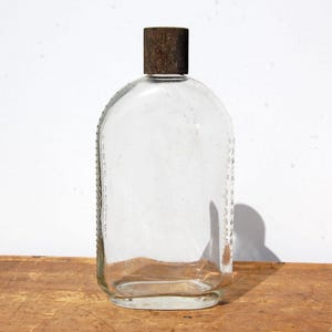 May include: A clear glass bottle with a rounded rectangular shape and a dark brown cap. The bottle has a textured surface and a series of vertical markings along the side. The cap appears to be made of a rough, possibly metal material.