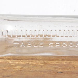 May include: A clear glass bottle with a dark metal cap. The bottle has measurements in tablespoons etched into the glass. The bottle is lying horizontally on a wooden surface.