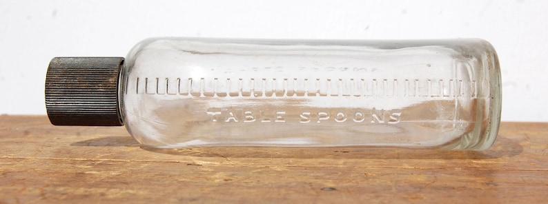 May include: A clear glass bottle with a dark metal cap. The bottle has measurements in tablespoons etched into the glass. The bottle is lying horizontally on a wooden surface.