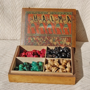 Puede incluir: Una caja de juego de madera con la palabra "HALMA" impresa en la tapa. La caja contiene cuatro compartimentos llenos de piezas de juego en rojo, negro, verde y beige.