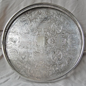 May include: A round silver-coloured serving tray featuring an intricate embossed floral and scroll pattern across its surface. The tray includes a raised pierced gallery rim, two side handles with shell-shaped accents, and a smooth central area.
