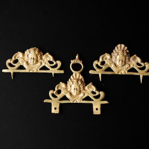 May include: Three antique gold-colored metal decorative pieces with cherub faces and ornate scrollwork. One piece has a ring, and the others have spikes or holes for mounting. The items are set against a black background.