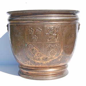 May include: An antique copper-coloured planter with a wide, rounded body and a decorative crest. The crest features floral and heraldic designs. The planter has a wide rim and a raised base, likely made of bronze.