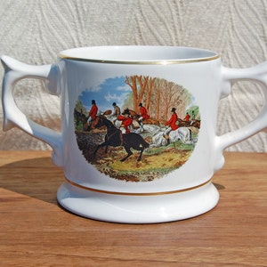 Puede incluir: Taza de cerámica blanca con dos asas. La taza presenta una ilustración colorida de personas a caballo cabalgando por un bosque.