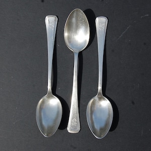 Puede incluir: Tres cucharas de metal plateado dispuestas verticalmente. Las dos cucharas exteriores tienen el cuenco hacia abajo, mientras que la central lo tiene hacia arriba. Cada mango presenta un detalle de monograma grabado en el extremo.