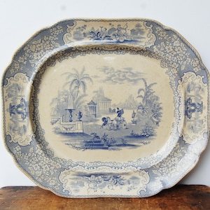 May include: An antique oval serving platter with a blue and white transferware design. The platter features a central landscape scene with figures, buildings, and trees, surrounded by a floral border. The edges have a scalloped design.