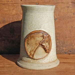 May include: A white ceramic mug with a brown horse head design. The mug has a speckled glaze and a rounded bottom.