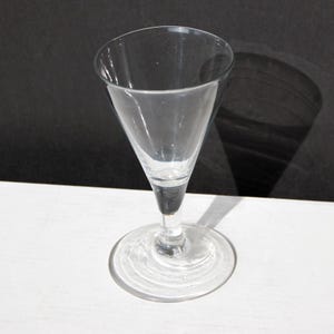 Könnte beinhalten: Ein klares Glas mit Stiel, konischer Schale und kreisförmigem Fuß. Das Glas hat einen langen, schlanken Stiel und eine weite Öffnung. Es besteht aus klarem Glas und dient zum Servieren von Getränken.