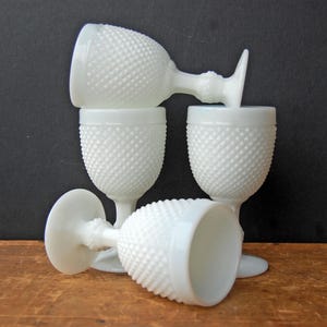 Puede incluir: Cuatro copas blancas con diseño de hobnail están dispuestas sobre una superficie de madera contra un fondo oscuro. Las copas están hechas de vidrio blanco opaco, con un patrón texturizado de puntos en relieve que cubre el cuenco y el tallo. Una copa está boca abajo.