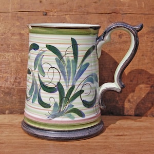 May include: A white ceramic beer stein with a green, blue and black floral design. The stein has a handle and a wide base.