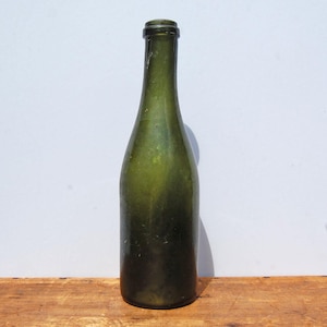 Op de afbeelding: Een hoge, cilindrische glazen fles met een diepe olijfgroene tint. De fles heeft een slanke hals, een afgeronde schouder en een dikke, verstevigde rand aan de bovenkant. Het glazen oppervlak vertoont subtiele variaties in kleur en textuur.