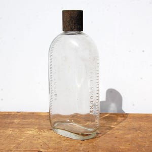 May include: Clear glass bottle with a brown cap. The bottle has a rectangular shape with rounded edges and a scale marked in tablespoons. The bottle is sitting on a wooden surface.