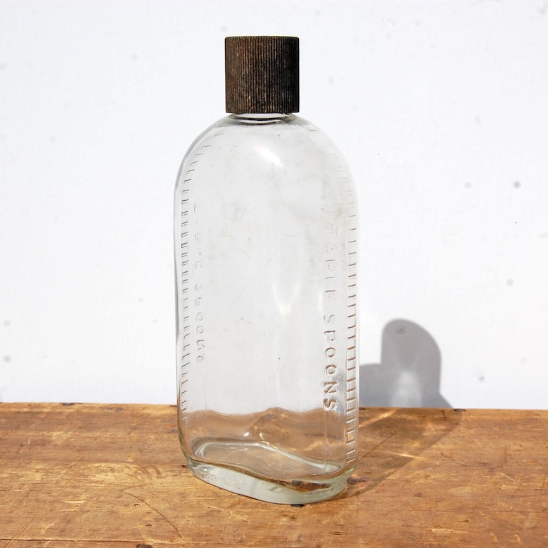 May include: Clear glass bottle with a brown cap. The bottle has a rectangular shape with rounded edges and a scale marked in tablespoons. The bottle is sitting on a wooden surface.
