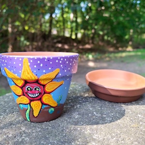 May include: A hand-painted terracotta flower pot with a matching saucer. The pot features a smiling sun design with yellow petals, a red face, and a purple sky background with white dots. The pot is approximately 4 inches tall.