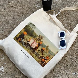 May include: Cream-colored tote bag with a vintage-style print featuring figures with pumpkin heads. The bag has the text "meet you there" printed on it. White sunglasses and a black cup are also visible.