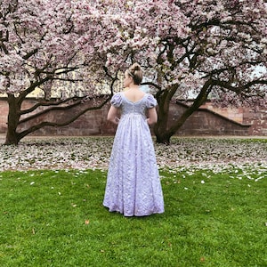 May include: A woman in a lavender gown stands in a grassy area with a magnolia tree behind her. The tree is in full bloom and petals are scattered on the ground.