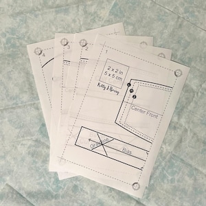 May include: A stack of white sewing pattern sheets with black printed diagrams and text. The text includes measurements of 2 x 2 inches and 5 x 5 cm, along with the name "Kitty J Berry". The patterns feature dotted lines, cutting guides, and labels like "Center Front" and "Bias".