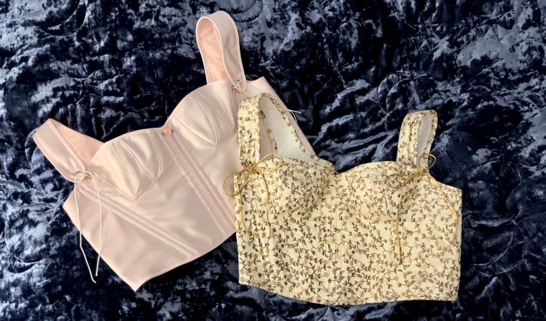 May include: Two corsets, one in a solid light pink satin and the other in a cream fabric with a floral pattern. Both corsets have a lace-up detail in the front and are on a blue velvet background.