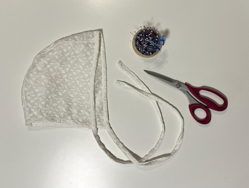 May include: A white bonnet with a leaf pattern, alongside a pincushion with pins and a pair of scissors with red handles. The bonnet has long ties. The items are arranged on a white surface.