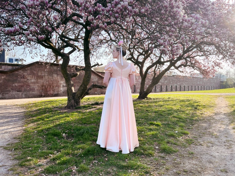 May include: A woman in a pale pink dress stands in a park with a magnolia tree in bloom behind her. The dress has a fitted bodice and a full skirt. The woman is wearing her hair up in a bun with a flower in it.
