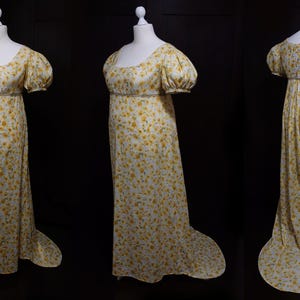 Op de afbeelding: Een witte jurk met een gele bloemenprint. De jurk heeft een getailleerd lijfje met een enkele dart, pofmouwen en een tweedelige rok met een sleep.