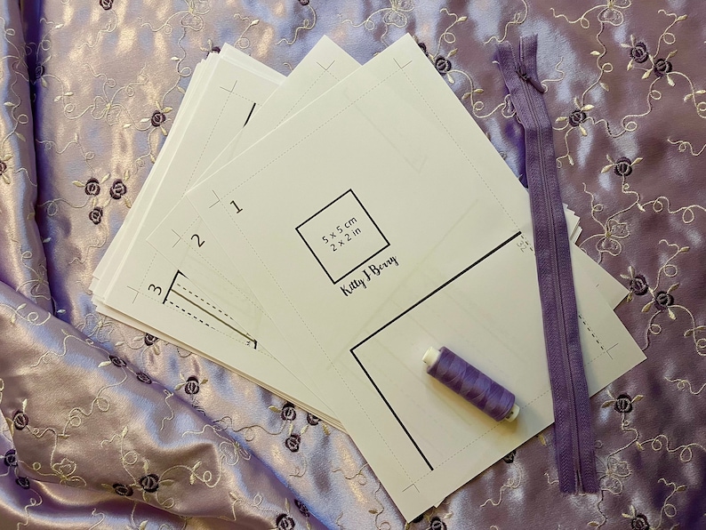 May include: A set of white paper patterns for a sewing project, with instructions for a 5 x 5 cm (2 x 2 in) square. The pattern is labeled "Kitty J Berry".  A purple zipper, purple thread, and a stack of additional patterns are also included.