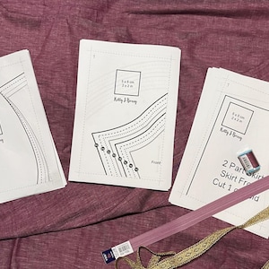 Op de afbeelding: Drie witte vellen papier met bedrukte naaipatronen. De patronen zijn voor een rok en zijn gelabeld "2 Part Skirt Front, Cut 1 of 1". Het papier ligt op een roze stofoppervlak.