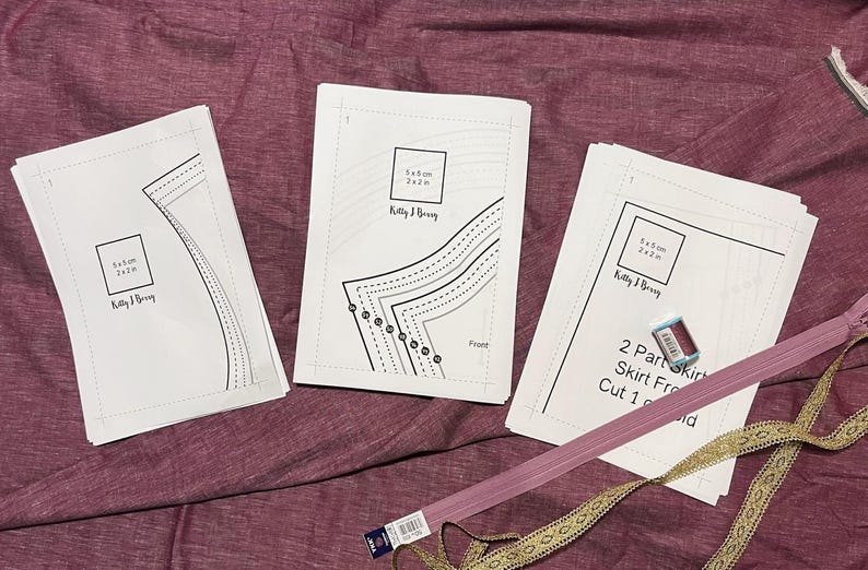 Op de afbeelding: Drie witte vellen papier met bedrukte naaipatronen. De patronen zijn voor een rok en zijn gelabeld "2 Part Skirt Front, Cut 1 of 1". Het papier ligt op een roze stofoppervlak.