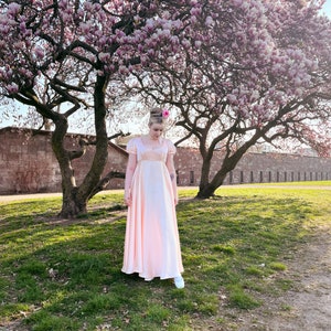 May include: A woman in a pink dress stands in front of a tree with pink blossoms. The tree is in front of a stone wall. The woman is wearing white shoes.