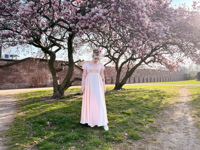 May include: A woman in a pink dress stands in front of a tree with pink blossoms. The tree is in front of a stone wall. The woman is wearing white shoes.
