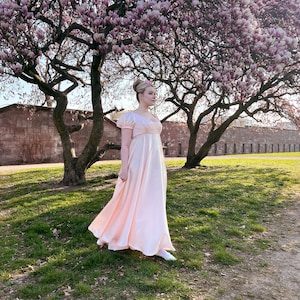 May include: A woman in a pale pink, empire waist dress stands in a grassy area with a large tree with pink blossoms in the background.