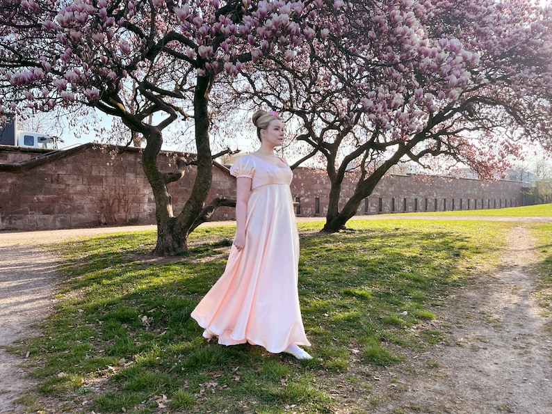 May include: A woman in a pale pink, empire waist dress stands in a grassy area with a large tree with pink blossoms in the background.