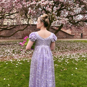 May include: A woman in a lavender dress with a floral pattern stands in a grassy area with pink flower petals scattered on the ground. She is holding a bouquet of pink tulips.