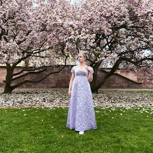 May include: A woman wearing a lavender gown stands in a grassy area with a large tree with pink blossoms in the background. The ground is covered in fallen petals.