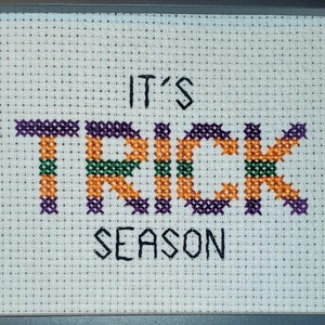May include: Cross-stitch artwork in a black frame. The text "IT'S TRICK SEASON" is stitched in orange, green, and purple thread on a white background. The artwork is a decorative piece, perfect for Halloween.