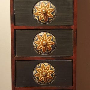 Puede incluir: Un pequeño mueble de madera con cinco cajones. El mueble está pintado de verde oscuro y rojo. Cada cajón tiene un pomo circular decorativo con un diseño de estrella dorada y azul.