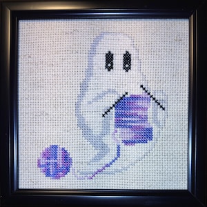 May include: Cross-stitch art of a white ghost knitting with black needles and purple yarn. The ghost has black eyes and is knitting a purple and blue scarf. A ball of purple and blue yarn sits on the bottom left. The artwork is framed in a black square frame.