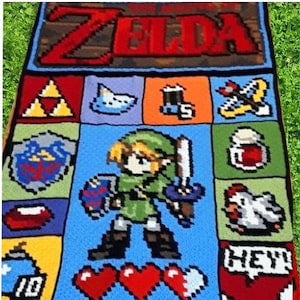 Könnte beinhalten: Eine gehäkelte Decke mit einem Pixel-Design des Videospielcharakters Link aus The Legend of Zelda. Die Decke ist blau, grün, rot, gelb und weiß. Die Decke enthält die Wörter "ZELDA" und "HEY!" in rot.