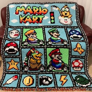 May include: A crocheted blanket featuring characters and items from the video game Mario Kart. The blanket is blue with black and white borders. The characters include Mario, Luigi, Toad, and Bowser. The items include a star, a mushroom, a banana, and a shell.