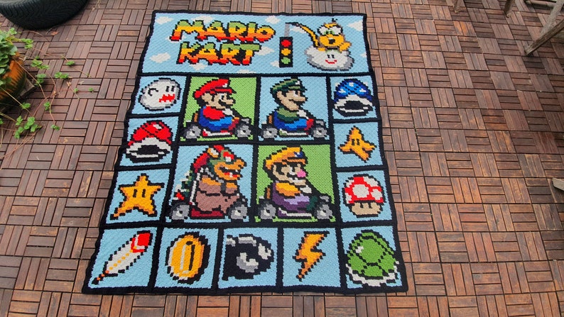 May include: A crocheted blanket featuring a pixelated design of characters from the video game Mario Kart. The blanket is blue with black outlines and features characters like Mario, Luigi, Bowser, and Toad.