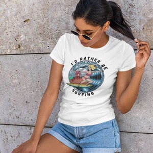 Puede incluir: Camiseta blanca con un gr&aacute;fico de una mujer surfeando en una ola con un volc&aacute;n en el fondo. El texto en la camiseta dice "I'd rather be surfing".
