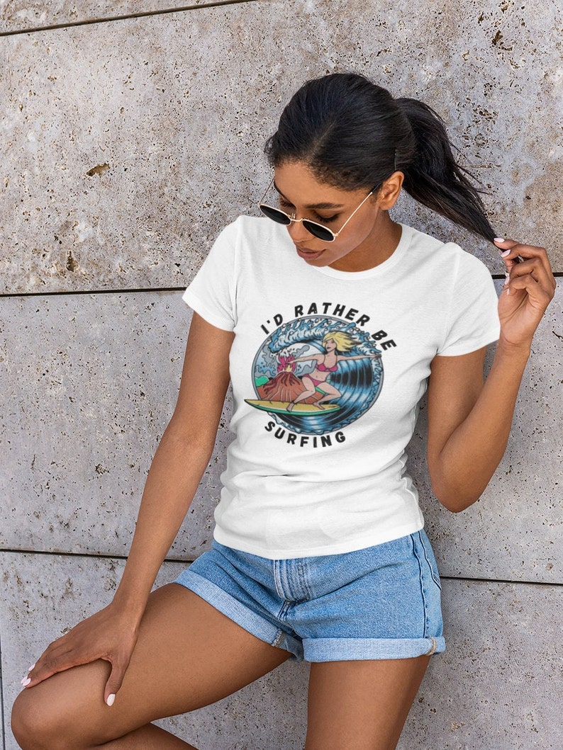 Puede incluir: Camiseta blanca con un gr&aacute;fico de una mujer surfeando en una ola con un volc&aacute;n en el fondo. El texto en la camiseta dice "I'd rather be surfing".
